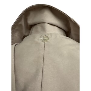 K&ouml;nnte beinhalten: Nahaufnahme eines beigefarbenen Jackenkragens mit Knopfverschluss. Der Kragen ist umgeklappt, und der Knopf ist ein kleiner, runder, klarer Knopf mit einem dekorativen Design. Die Jacke scheint aus einem glatten Stoff zu sein.