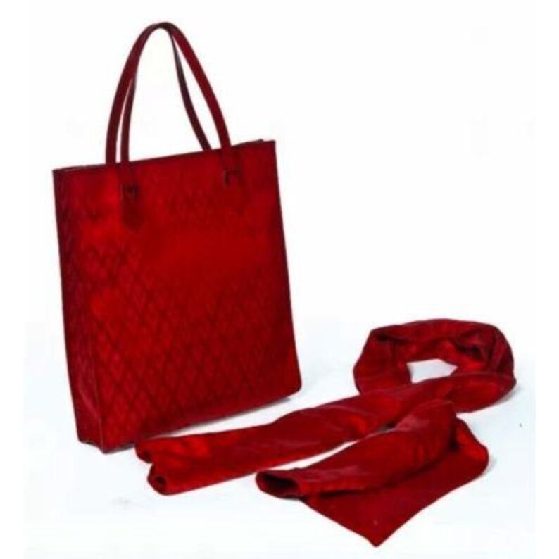 May include: A red tote bag with matching handles and a diamond pattern, alongside a long, red scarf. The bag is rectangular and stands upright. The scarf is folded and draped on a white surface.