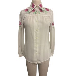 Op de afbeelding: Een witte blouse met lange mouwen, een puntkraag en een knoopsluiting. De kraag en mouwen zijn versierd met geborduurde roze rozen en groene bladeren. De blouse is gemaakt van een lichte, semi-transparante stof.