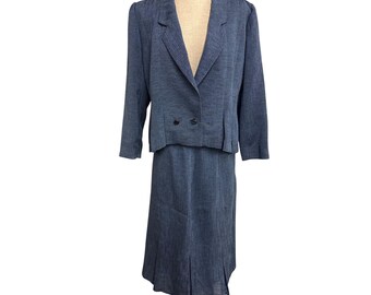 May include: A two-piece denim blue suit, consisting of a blazer and a knee-length skirt. The blazer has a notched collar, two buttons, and long sleeves. The skirt has a pleated design. The suit is displayed on a mannequin.