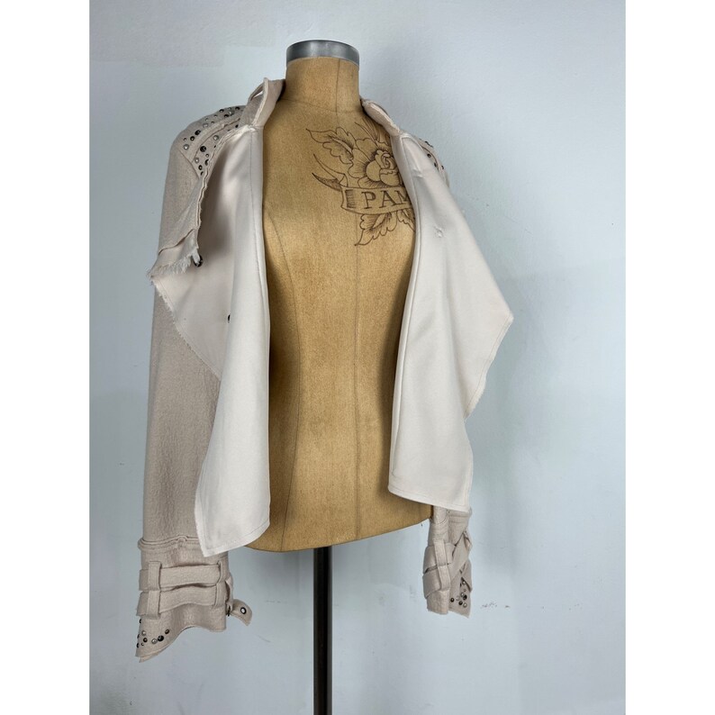 May include: Beige jacket with silver studs on the shoulders and cuffs. The jacket has a draped design with an open front, revealing a white lining. The cuffs feature buckle details. The jacket is displayed on a mannequin.