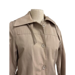 K&ouml;nnte beinhalten: Ein beigefarbener Trenchcoat mit klassischem Design. Der Mantel hat einen Reverskragen, Knopfverschl&uuml;sse und einen Tailleng&uuml;rtel. Der Mantel ist aus strapazierf&auml;higem Stoff gefertigt und f&uuml;r verschiedene Wetterbedingungen geeignet.