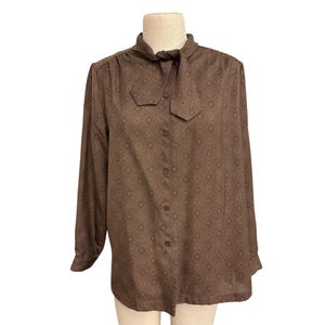 Pode incluir: Uma blusa marrom de manga comprida com gola de laço. A blusa apresenta um padrão geométrico repetitivo em um tom mais escuro de marrom. O tecido parece leve e a blusa é abotoada na frente.