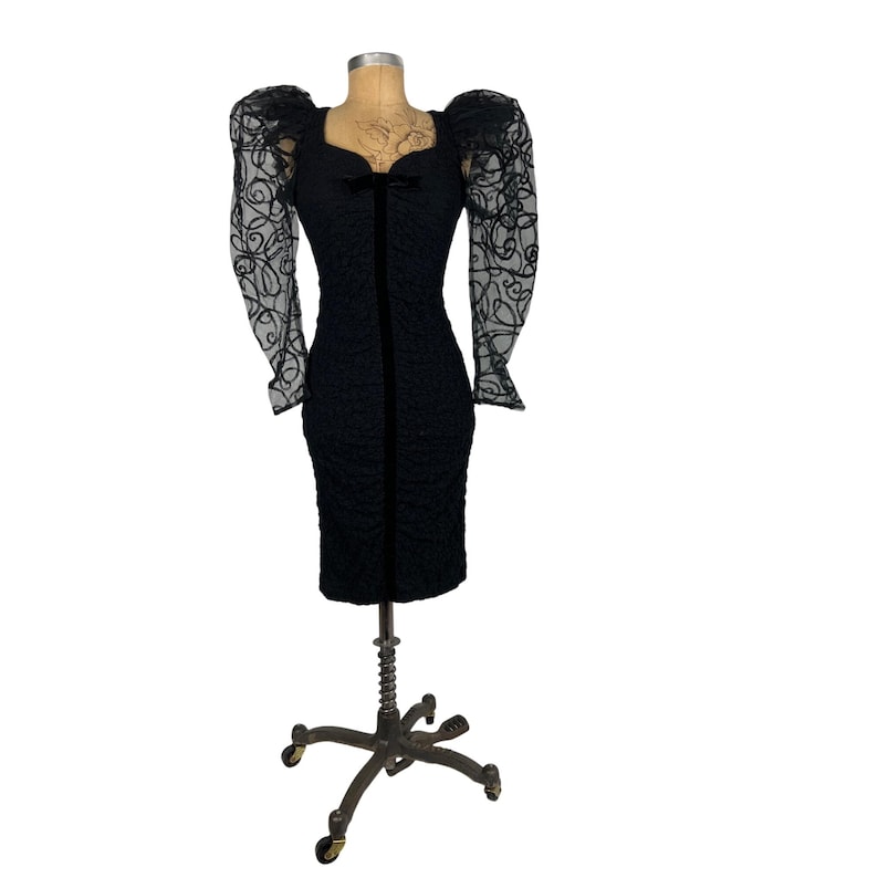 May include: A black dress with a sweetheart neckline and puff sleeves. The sleeves are made of sheer fabric with a floral pattern. The dress is fitted and has a midi length.