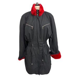 Puede incluir: Abrigo negro con cuello y puños de piel sintética roja. El abrigo tiene cintura con cordón, dos bolsillos en el pecho con cremallera y dos bolsillos inferiores. El cuello tiene tres botones. El abrigo se muestra en un maniquí.