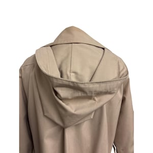 K&ouml;nnte beinhalten: Ein beigefarbener Mantel mit Kapuze, von hinten gesehen, mit hochgezogener Kapuze. Der Mantel ist einfarbig, und die Kapuze ist ein herausragendes Merkmal. Der Stoff scheint ein mittelschweres Material zu sein.