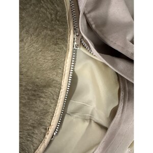 K&ouml;nnte beinhalten: Nahaufnahme einer beigefarbenen Jacke mit silbernem Rei&szlig;verschluss. Das Innenfutter der Jacke ist hellbeige, und das Au&szlig;enmaterial scheint ein glatter, gewebter Stoff zu sein. Die linke Seite der Jacke hat ein flauschiges, olivgr&uuml;nes Material.