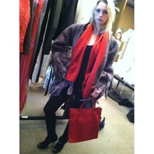 May include: A woman wearing a gray coat with a red scarf and holding a red tote bag. The tote bag has a diamond pattern. The woman is wearing black leggings and black heels.