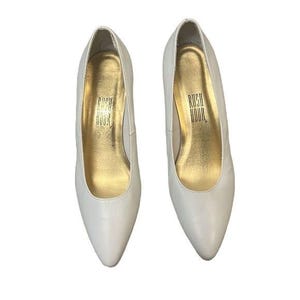 May include: A pair of white leather pumps with pointed toes. The interior of each shoe is gold with the text "RUSH HOUR". These classic heels are a timeless addition to any wardrobe.