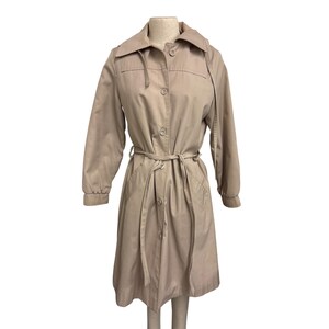 K&ouml;nnte beinhalten: Ein beigefarbener Trenchcoat mit Knopfleiste, passendem G&uuml;rtel und Kragen. Der Mantel hat lange &Auml;rmel und wird auf einer Schaufensterpuppe pr&auml;sentiert. Die Farbe des Mantels ist neutral.