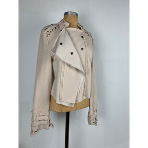May include: A light beige jacket with a unique asymmetrical design. The jacket features silver-colored studs on the shoulders and cuffs, with multiple button closures and decorative straps on the sleeves. The collar is a stand-up style.