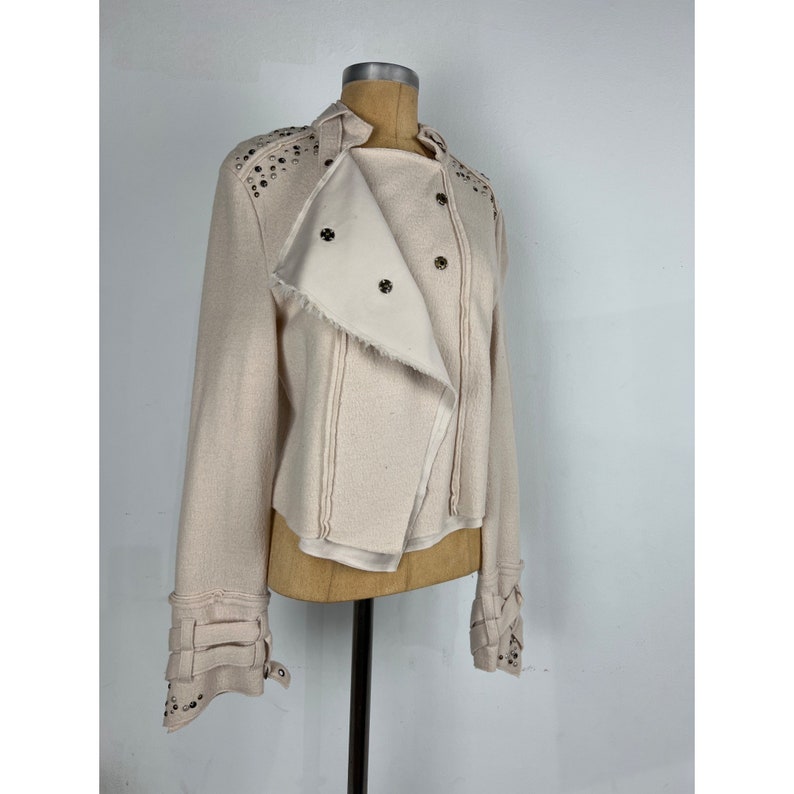 May include: A light beige jacket with a unique asymmetrical design. The jacket features silver-colored studs on the shoulders and cuffs, with multiple button closures and decorative straps on the sleeves. The collar is a stand-up style.