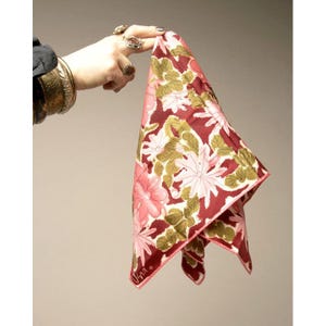 May include: A floral patterned scarf in shades of burgundy, pink, white, and olive green. The scarf is held up, displaying its square shape and the intricate flower design. The scarf has a pink border and the signature "Una" is visible.
