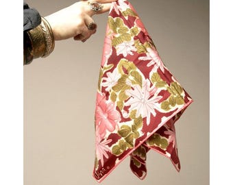 May include: A square scarf with a floral pattern in burgundy, pink, white, and olive green. The scarf is held up, showing its design and shape. It has a pink border and the signature "Una" is visible in the corner.