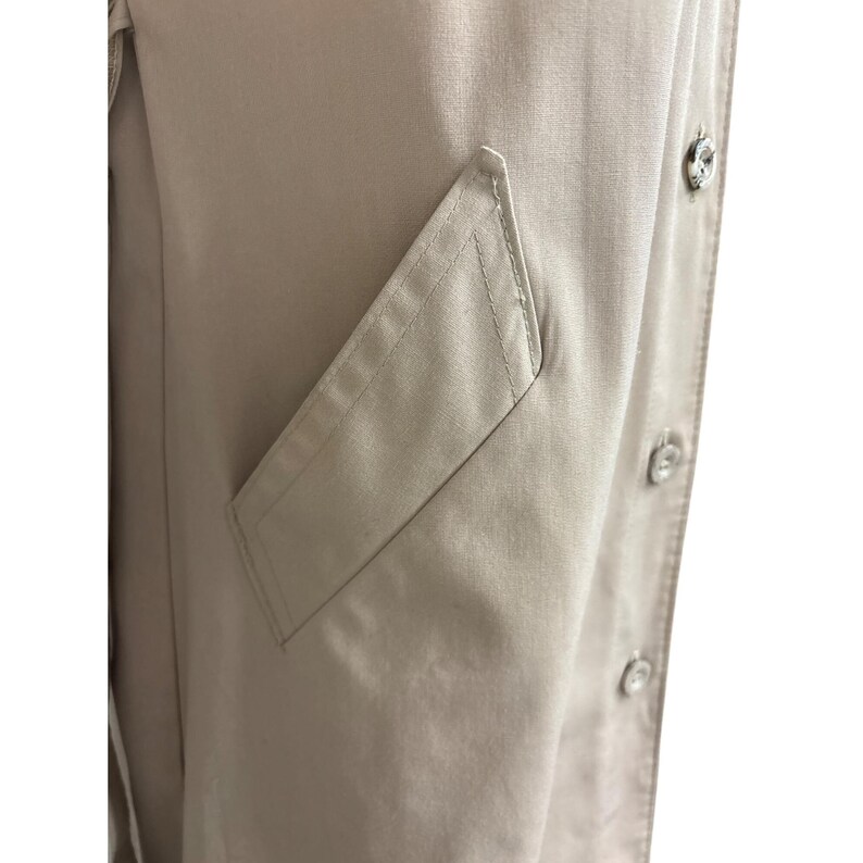 K&ouml;nnte beinhalten: Nahaufnahme eines beigefarbenen Trenchcoats. Der Mantel hat eine Klappentasche und dekorative Kn&ouml;pfe. Der Stoff scheint ein glattes, gewebtes Material zu sein. Der Mantel hat eine neutrale Farbe und ist f&uuml;r verschiedene Outfits geeignet.