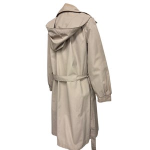 K&ouml;nnte beinhalten: Beigefarbener Trenchcoat mit Kapuze, G&uuml;rtel und langen &Auml;rmeln. Der Mantel ist aus einem glatten Stoff gefertigt und hat ein klassisches Design. Der Mantel ist neutralfarben und f&uuml;r verschiedene Outfits geeignet.