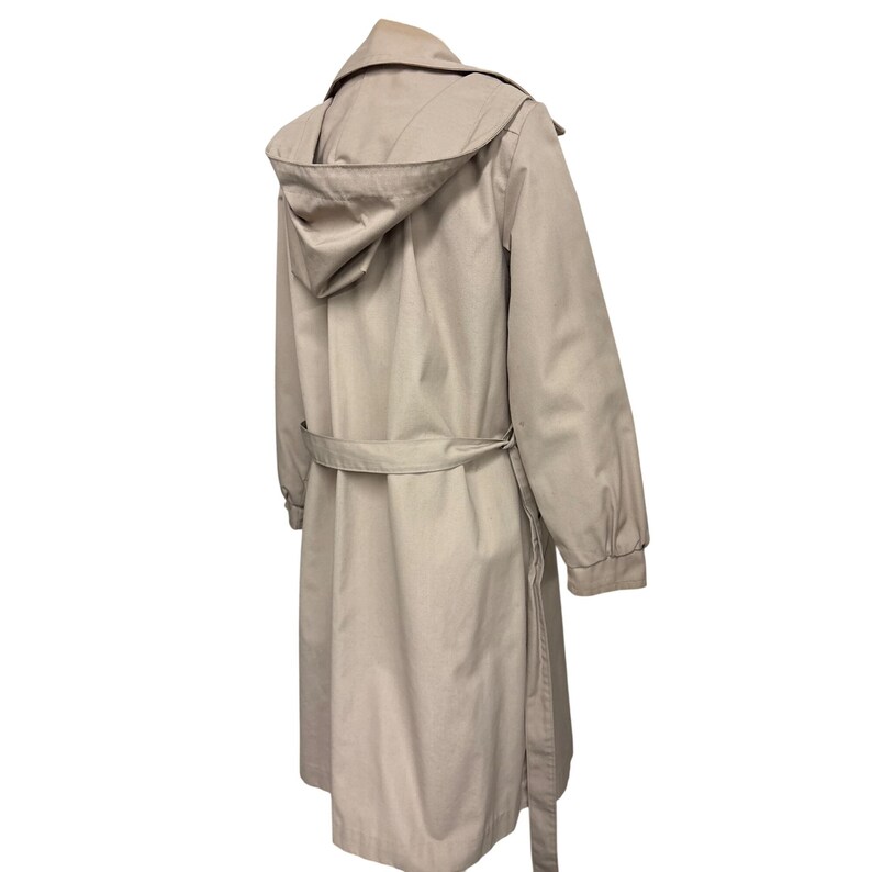 K&ouml;nnte beinhalten: Beigefarbener Trenchcoat mit Kapuze, G&uuml;rtel und langen &Auml;rmeln. Der Mantel ist aus einem glatten Stoff gefertigt und hat ein klassisches Design. Der Mantel ist neutralfarben und f&uuml;r verschiedene Outfits geeignet.