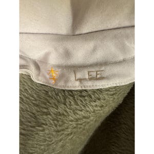 K&ouml;nnte beinhalten: Nahaufnahme eines hellgrauen Stoffes mit dem gestickten Wort "LEE" in passender Farbe. Links neben dem Text ist ein goldfarbenes Symbol gestickt. Der Stoff ist vor einem verschwommenen, olivgr&uuml;nen Hintergrund platziert.