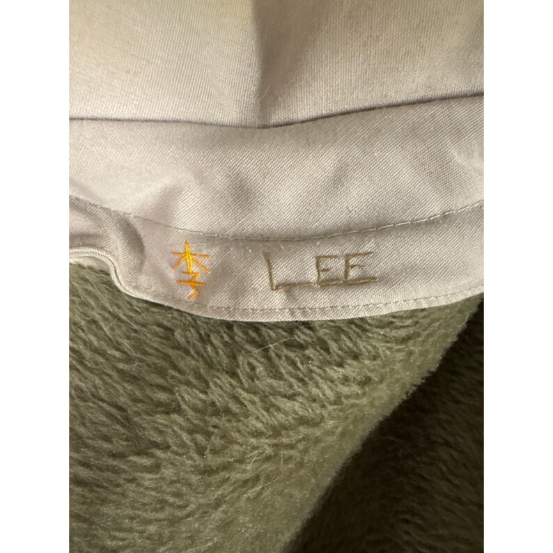 K&ouml;nnte beinhalten: Nahaufnahme eines hellgrauen Stoffes mit dem gestickten Wort "LEE" in passender Farbe. Links neben dem Text ist ein goldfarbenes Symbol gestickt. Der Stoff ist vor einem verschwommenen, olivgr&uuml;nen Hintergrund platziert.