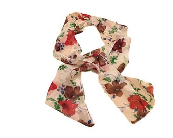 May include: A cream-coloured scarf with a floral pattern. The scarf features red and brown flowers, green leaves, and purple accents. The scarf is made of a lightweight material.