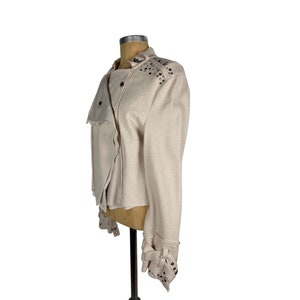 May include: A light beige jacket with a textured fabric. The jacket features a stand-up collar, asymmetrical front, and silver-tone stud embellishments on the shoulders and cuffs. The cuffs have decorative straps.