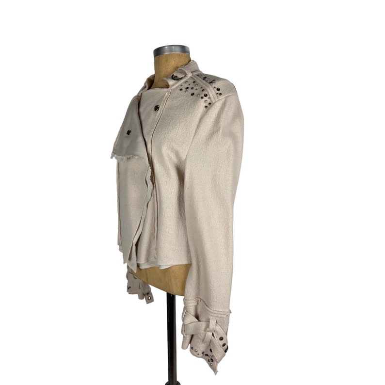 May include: A light beige jacket with a textured fabric. The jacket features a stand-up collar, asymmetrical front, and silver-tone stud embellishments on the shoulders and cuffs. The cuffs have decorative straps.