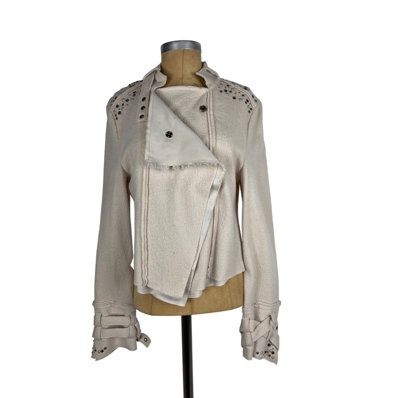 May include: Cream-colored jacket with an asymmetrical design. The jacket features a notched collar, long sleeves, and decorative metal studs along the shoulders and cuffs. The jacket is displayed on a mannequin.