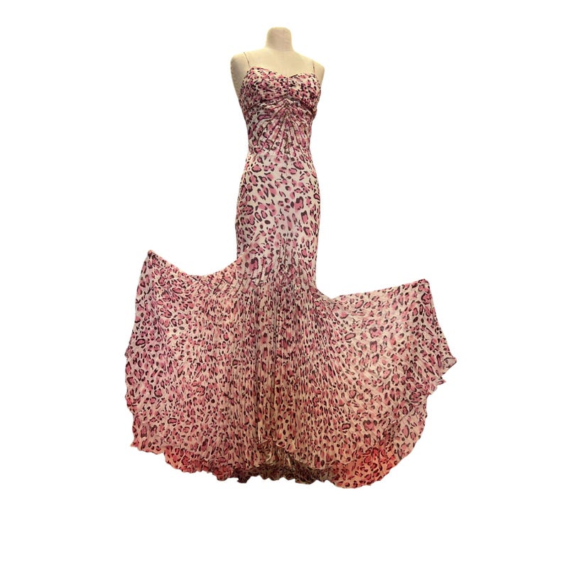 May include: A floor-length, strapless gown on a mannequin. The dress features a pink and white leopard print pattern. The bodice is ruched, and the skirt flares out into a flowing, pleated design. The dress is made of a lightweight fabric.