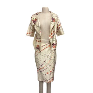 May include: A two-piece set consisting of a short-sleeved jacket and a knee-length skirt. Both pieces are made of a cream-colored fabric featuring a repeating diagonal lattice pattern of small pink, orange, and purple floral clusters throughout.