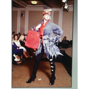 May include: A model on a runway wearing a unique ensemble. The outfit includes a gray, feathery coat, black leggings with white polka dots, and black heels. A red scarf and handbag add pops of color. The model also wears a patterned hat.
