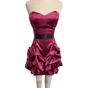 Puede incluir: Un vestido corto sin tirantes en color magenta intenso. El vestido presenta un escote corazón, un cinturón de satén negro y una falda con volantes. El vestido se exhibe en un maniquí.