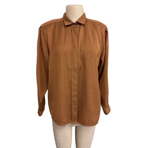 May include: A long-sleeved, button-down shirt in a warm brown colour. The shirt features a classic collar, shoulder pads, and a tailored fit. The fabric appears to have a subtle texture, adding depth to the solid colour. The shirt is displayed on a mannequin.