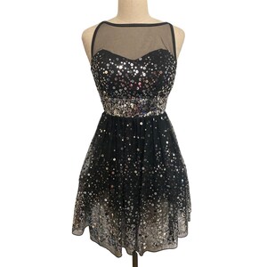 May include: A black sequined party dress with a sweetheart neckline and a sheer overlay. The dress features a fitted bodice and a flared skirt, with silver sequins adorning the bodice and waistline. The dress is displayed on a mannequin.