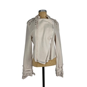 May include: Cream-colored asymmetrical jacket with long sleeves. The jacket features decorative studs on the shoulders and cuffs, along with multiple straps and buckles. The design includes a wrap-style front and a high collar.
