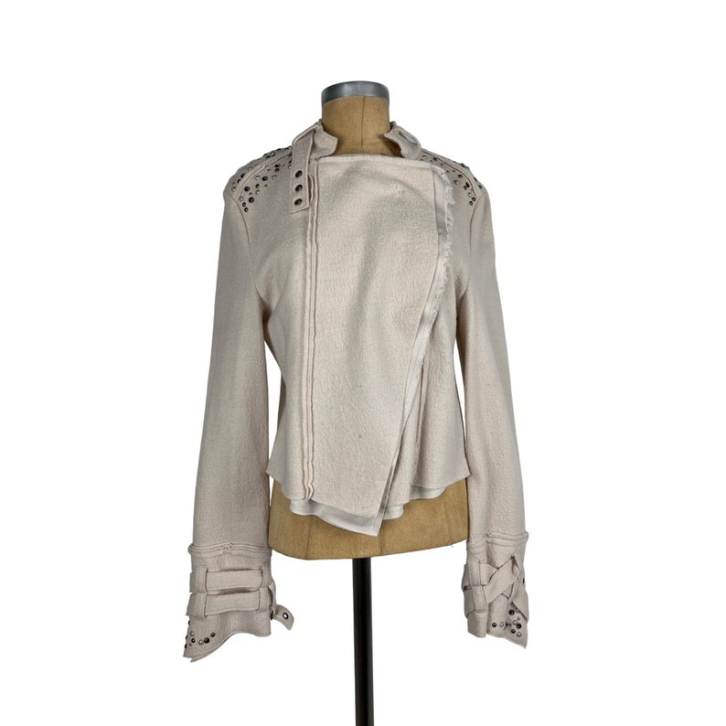 May include: Cream-colored asymmetrical jacket with long sleeves. The jacket features decorative studs on the shoulders and cuffs, along with multiple straps and buckles. The design includes a wrap-style front and a high collar.