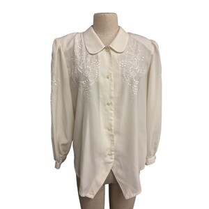 May include: A cream-coloured blouse with long sleeves and a rounded collar. The blouse features intricate white floral embroidery across the chest and sleeves. The button-down shirt is displayed on a mannequin.