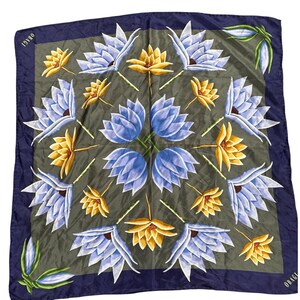 Può includere: Foulard quadrato in seta con motivo floreale. La sciarpa presenta un bordo blu scuro e un motivo centrale di fiori blu e gialli su sfondo verde. La parola "OBAGI" è stampata sul bordo.