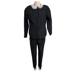 May include: A black quilted suit with a white lace collar. The suit includes a long-sleeved jacket and matching trousers. The jacket has a button closure, and the trousers have a straight leg design. The suit is displayed on a mannequin.