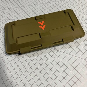 以下が含まれることがあります： オリーブグリーンの長方形の容器で、ヒンジ付きの蓋と2つのオレンジ色の下向きの矢印が付いています。容器はテクスチャ加工された表面で、おそらくプラスチック製です。
