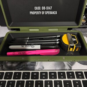 以下が含まれることがあります： 「CASE: DB-S147 PROPERTY OF SPERANZA」の文字が入った、開いたオリーブグリーンのケース。ケースの中には黒いペン、白いSharpie、ピンクの蛍光ペン、黄と黒のメジャーが入っています。ノートパソコンのキーボードが手前にあります。