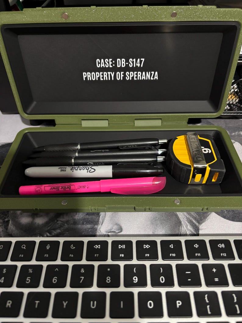 以下が含まれることがあります： 「CASE: DB-S147 PROPERTY OF SPERANZA」の文字が入った、開いたオリーブグリーンのケース。ケースの中には黒いペン、白いSharpie、ピンクの蛍光ペン、黄と黒のメジャーが入っています。ノートパソコンのキーボードが手前にあります。