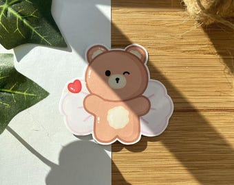 May include: A die-cut sticker featuring a cartoon bear with a winking expression. The bear is light brown with a white belly and is set against a pink and white cloud design. A small red heart is also included in the design.