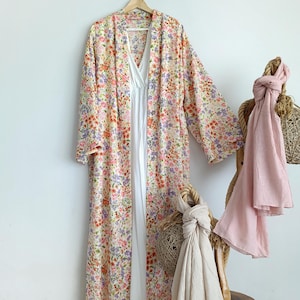 Peut inclure: Une longue robe kimono à imprimé floral avec une robe blanche en dessous est suspendue à un cintre en bois. La robe présente un fond crème avec des fleurs roses, violettes et orange. Une écharpe rose clair et une écharpe beige sont drapées sur un support en bois.