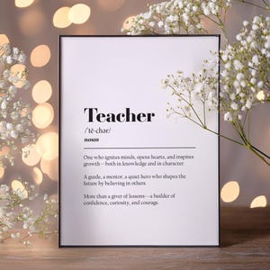 May include: A framed print with the definition of "Teacher" in black text on a white background. The definition includes the phonetic pronunciation and a description of a teacher's role. White flowers and bokeh lights are in the background.