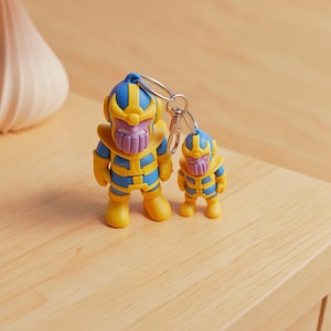 May include: A keychain featuring two stylized figures of a purple-skinned character with a gold and blue helmet and armor. The larger figure is attached to a silver metal split ring and lobster clasp, while the smaller figure is attached to the larger one.