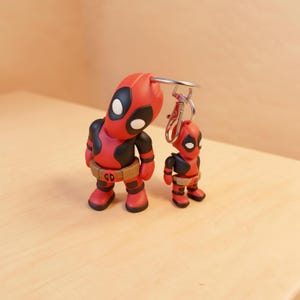 May include: A keychain featuring two Deadpool figures in red and black suits with white eye patches and brown utility belts. The larger figure is attached to a silver metal split ring and lobster clasp, while the smaller figure hangs beside it.