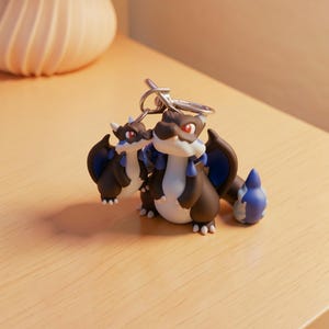 May include: Two dragon-shaped keychains. The dragons are primarily brown with blue wings and accents, white bellies, and red eyes. The keychains are made of a glossy material and are attached to a silver metal ring.