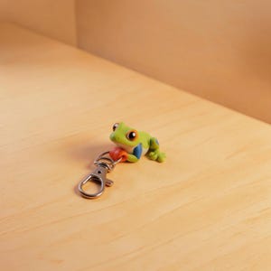 May include: A small, sculpted green frog figurine with orange eyes and blue side markings, attached to a silver-toned metal lobster clasp keychain. The frog is depicted in a resting position with its front limbs tucked near its chin.