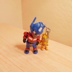 May include: A blue, red, and grey robot figurine keychain featuring a larger character with a helmet and a smaller yellow character attached by a silver metal lobster clasp. The figures have a smooth, matte finish and stylized, rounded proportions.