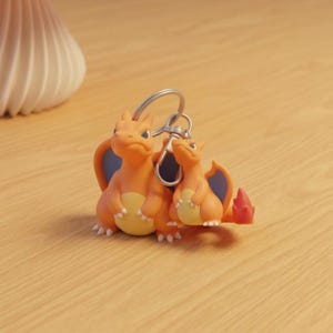 May include: Orange and yellow Charizard keychain featuring two figures with blue wings and red flames. The keychain is attached to a silver metal ring. The figures are on a wooden surface.